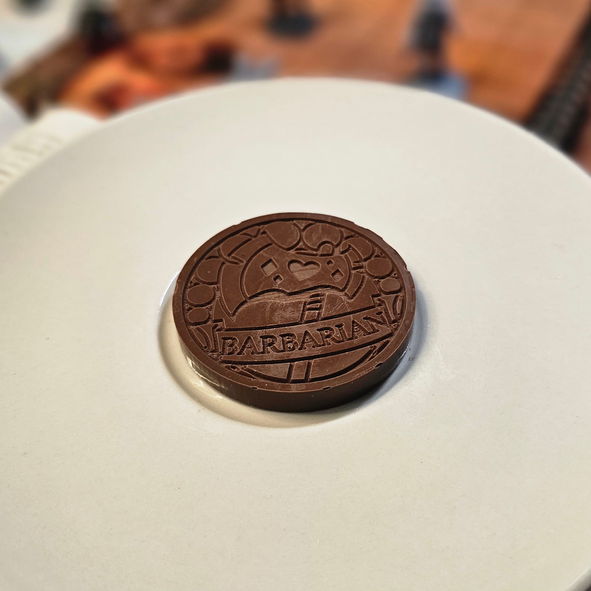 Home Milk chocolate crunch barbarian inspiration coin displayed on a white plate with tabletop RPG miniatures, dice, and dungeon playmat blurred in the background.