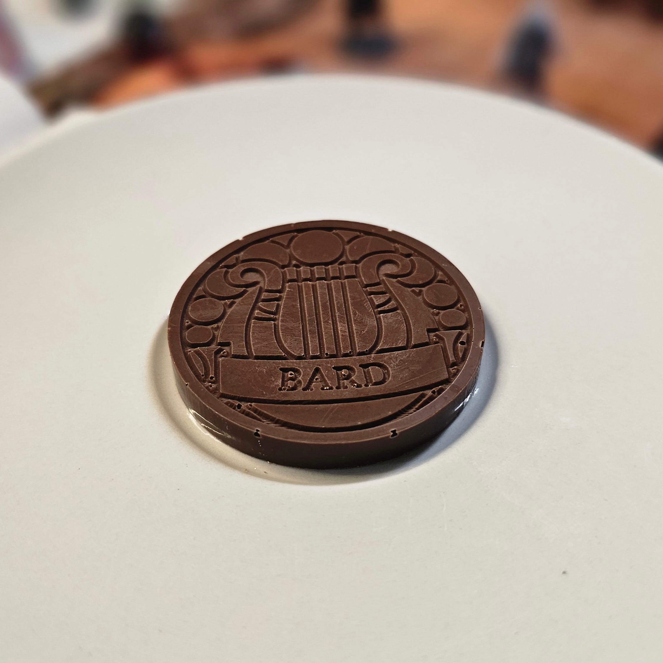 Home Milk chocolate crunch bard inspiration coin displayed on a white plate with tabletop RPG miniatures, dice, and dungeon playmat blurred in the background.