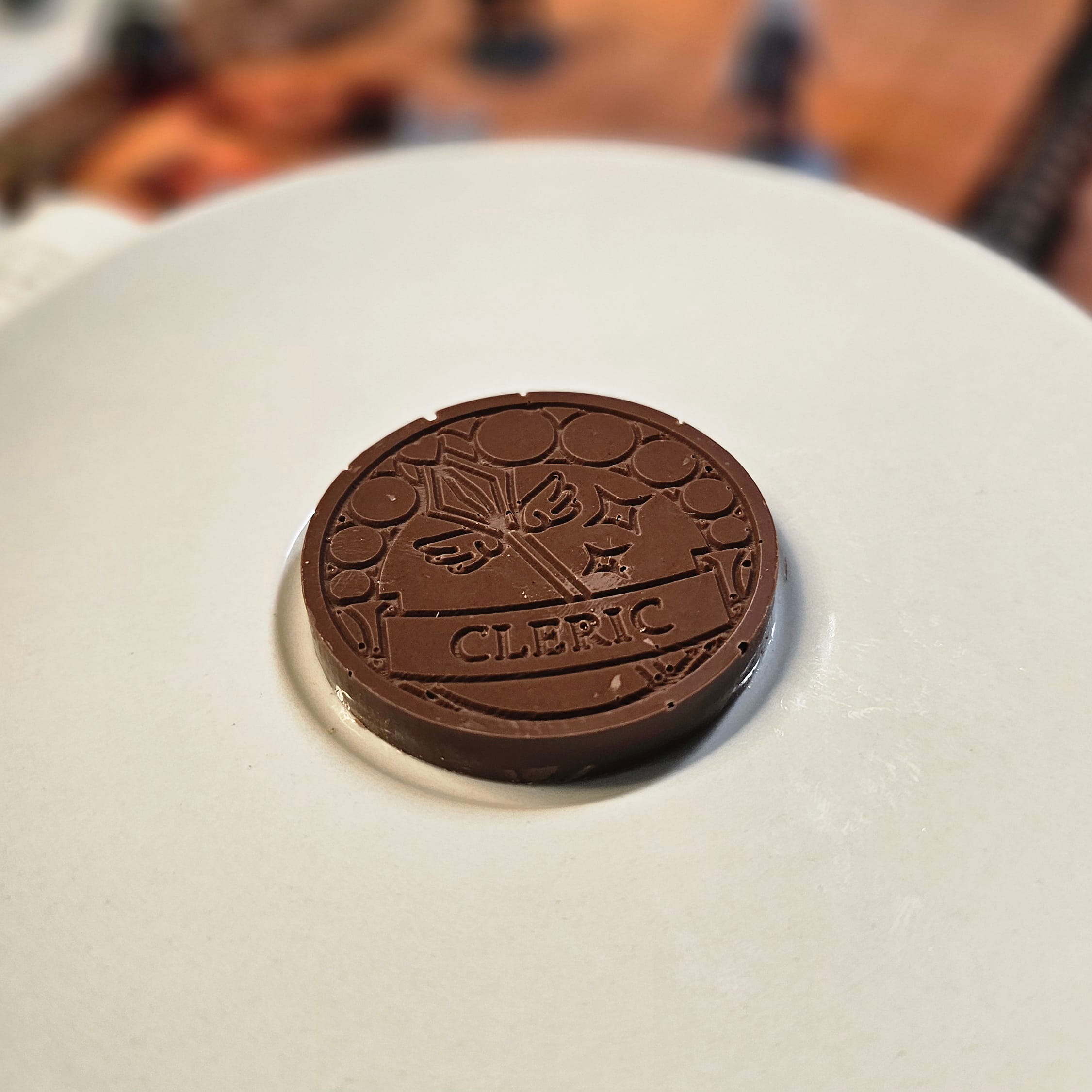 Home Milk chocolate crunch cleric inspiration coin displayed on a white plate with tabletop RPG miniatures, dice, and dungeon playmat blurred in the background.
