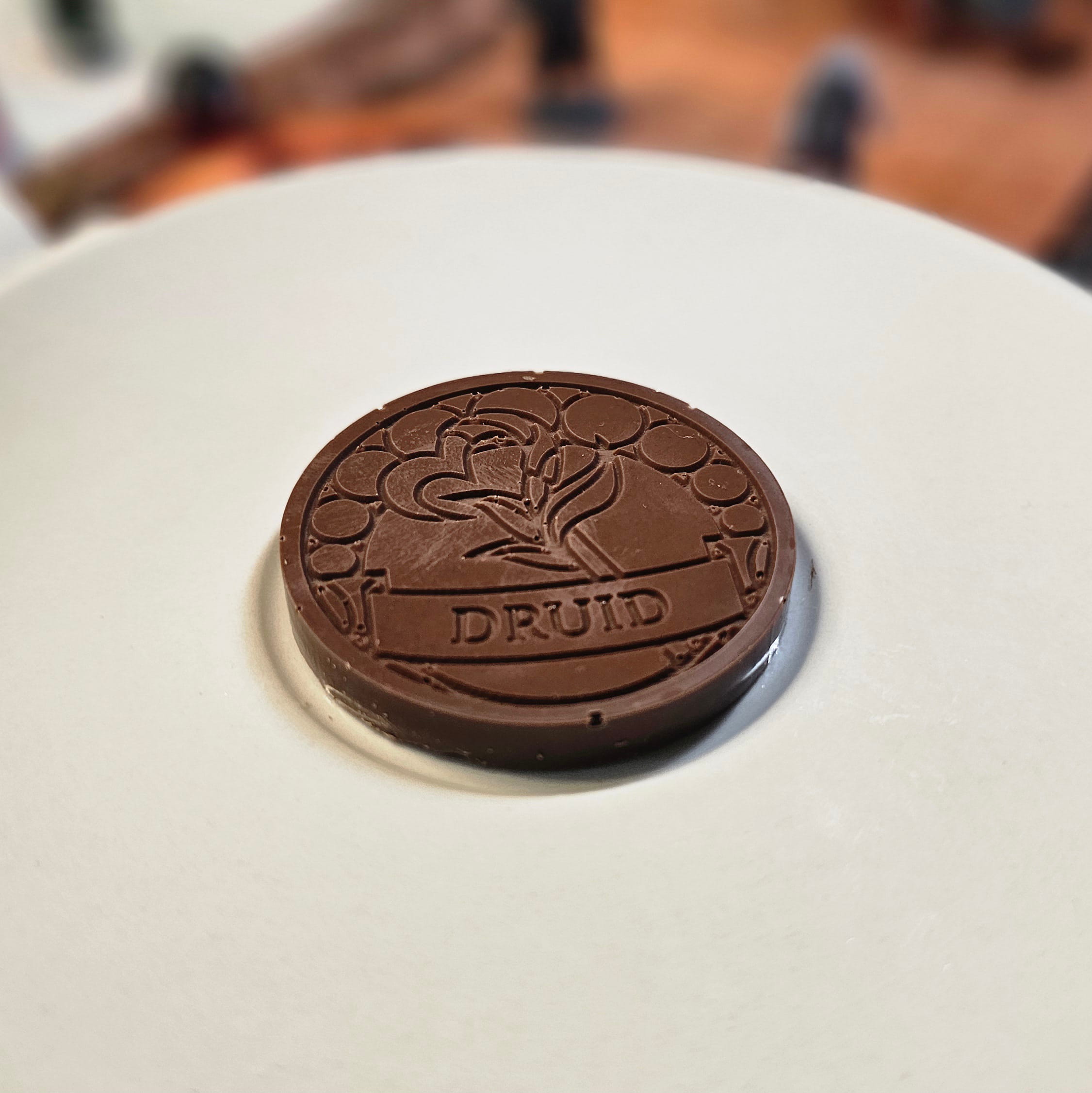 Home Milk chocolate crunch druid inspiration coin displayed on a white plate with tabletop RPG miniatures, dice, and dungeon playmat blurred in the background.