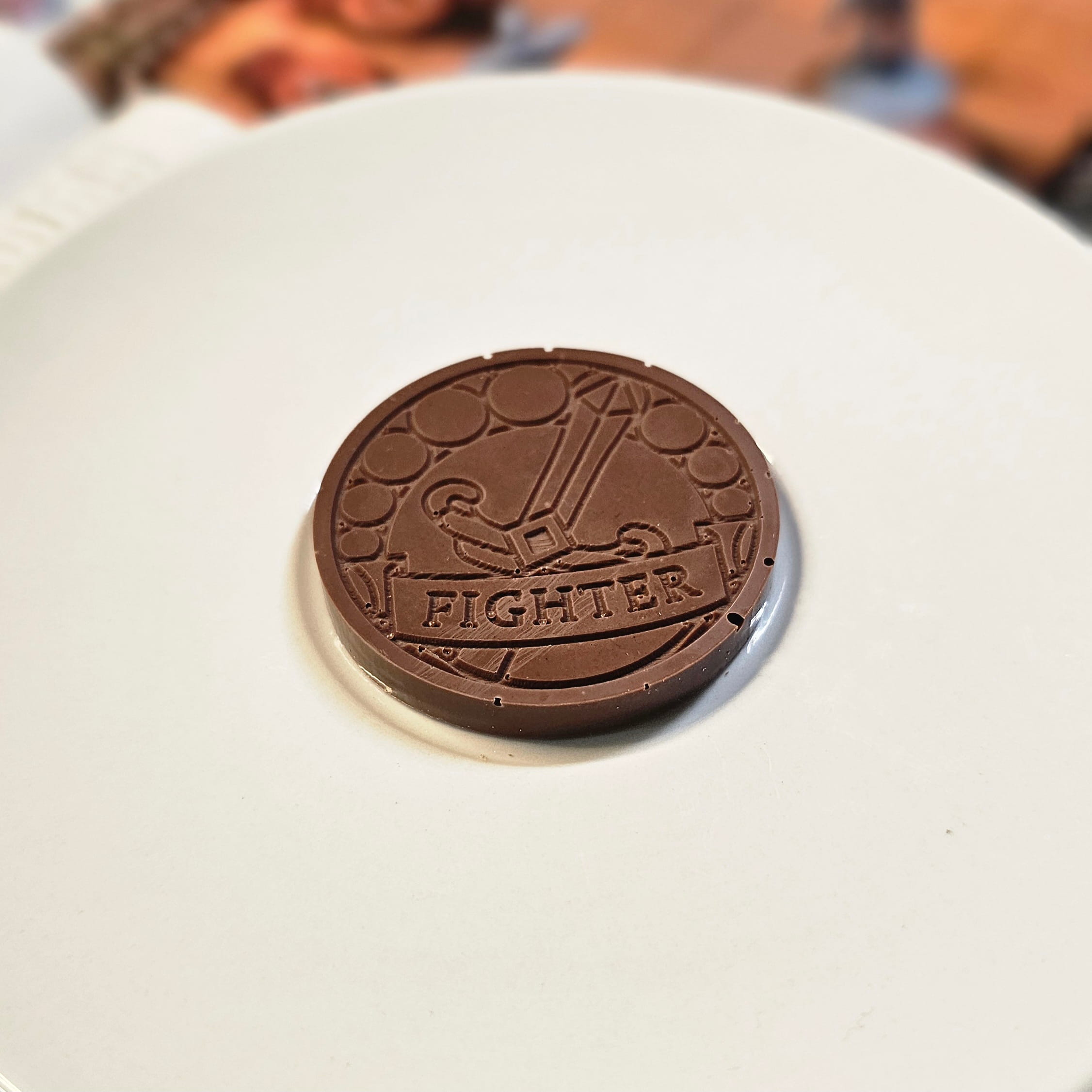 Home Milk chocolate crunch fighter inspiration coin displayed on a white plate with tabletop RPG miniatures, dice, and dungeon playmat blurred in the background.