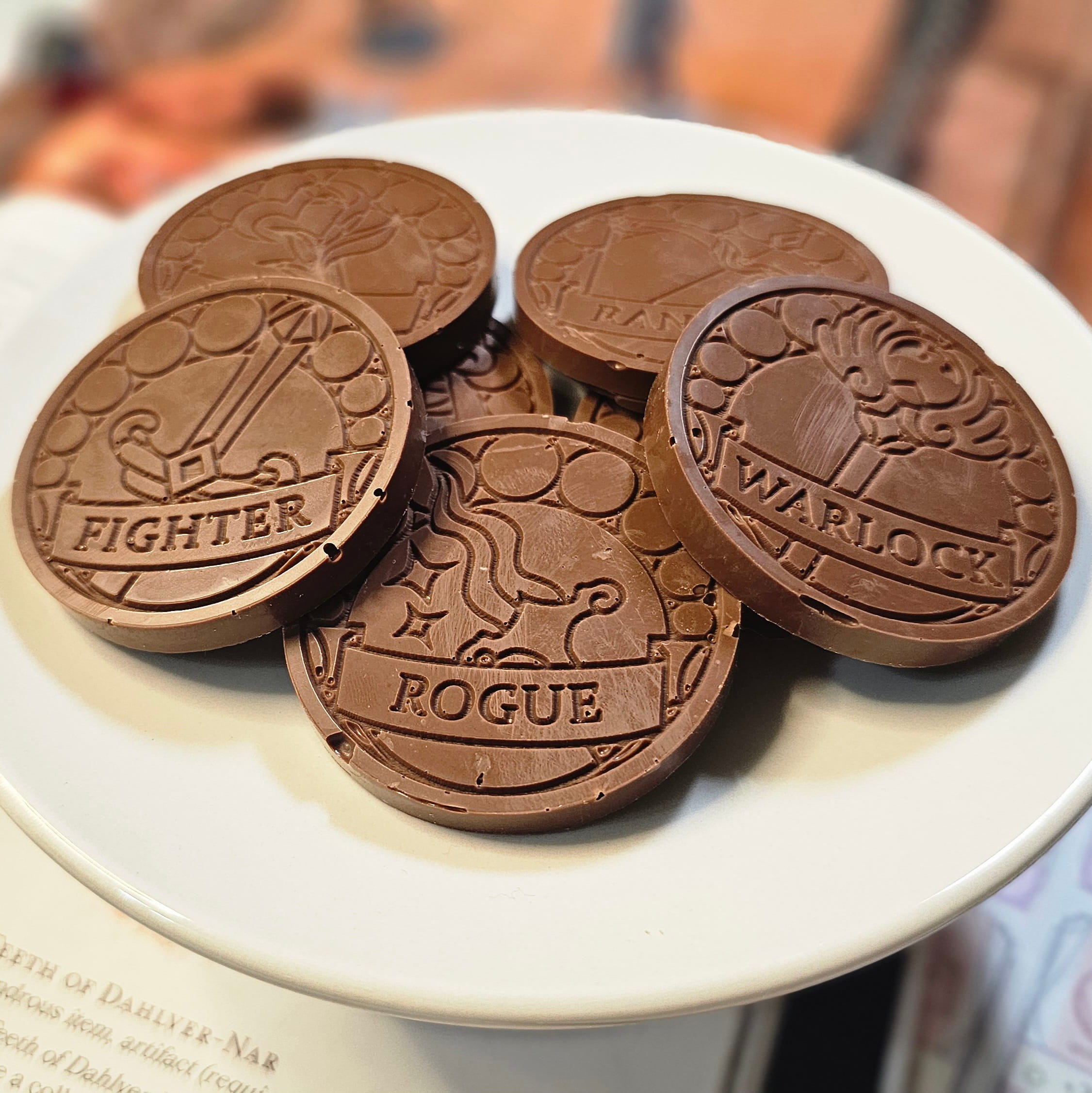 Home Milk chocolate crunch D&D inspiration coins displayed on a white plate with tabletop RPG miniatures, dice, character sheet, D&D player handbook, and dungeon playmat blurred in the background.