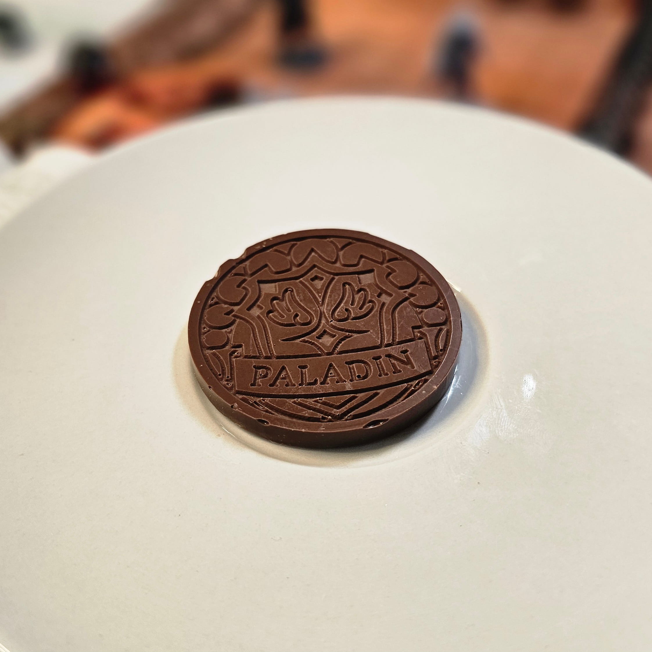 Home Milk chocolate crunch paladin inspiration coin displayed on a white plate with tabletop RPG miniatures, dice, and dungeon playmat blurred in the background.