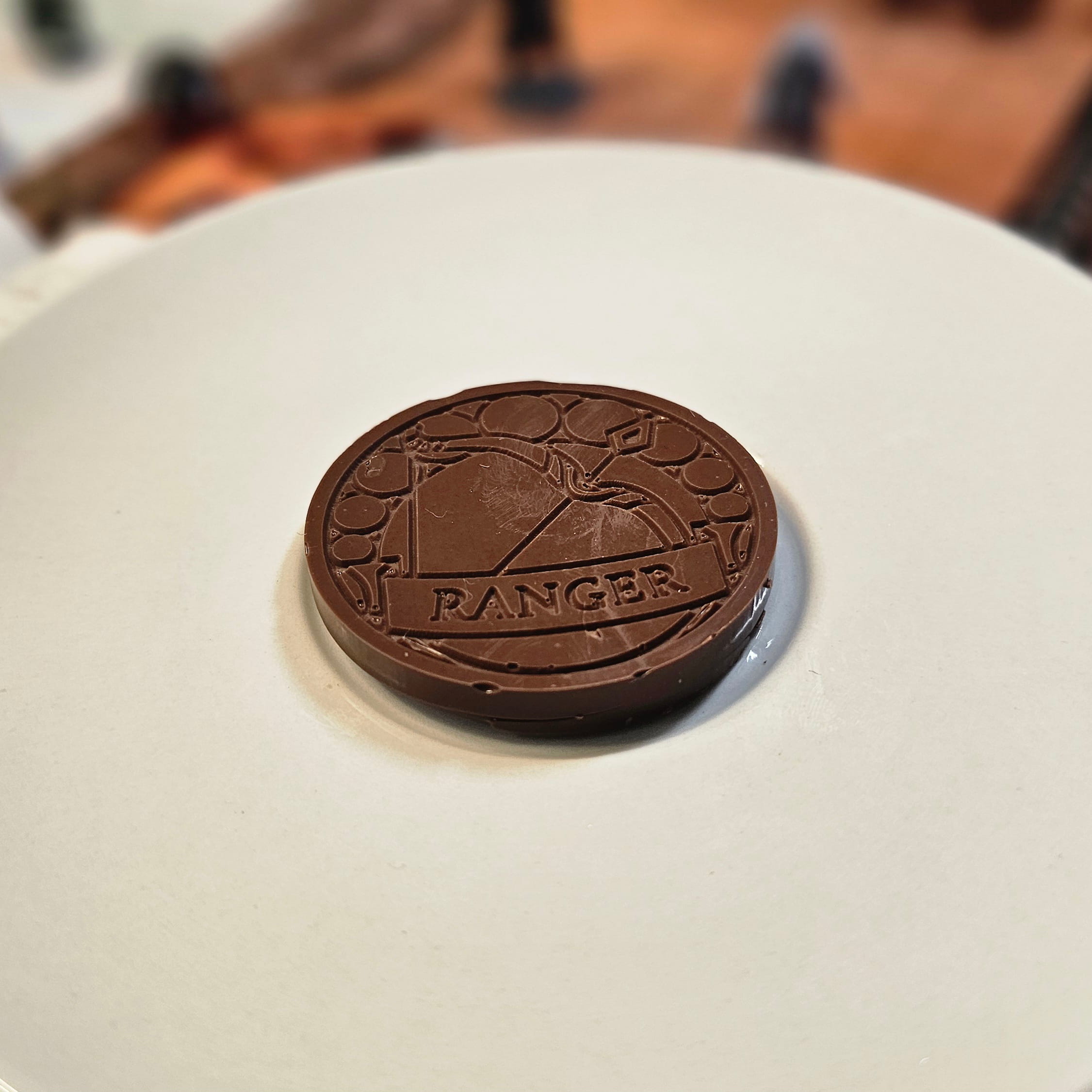 Home Milk chocolate crunch ranger inspiration coin displayed on a white plate with tabletop RPG miniatures, dice, and dungeon playmat blurred in the background.