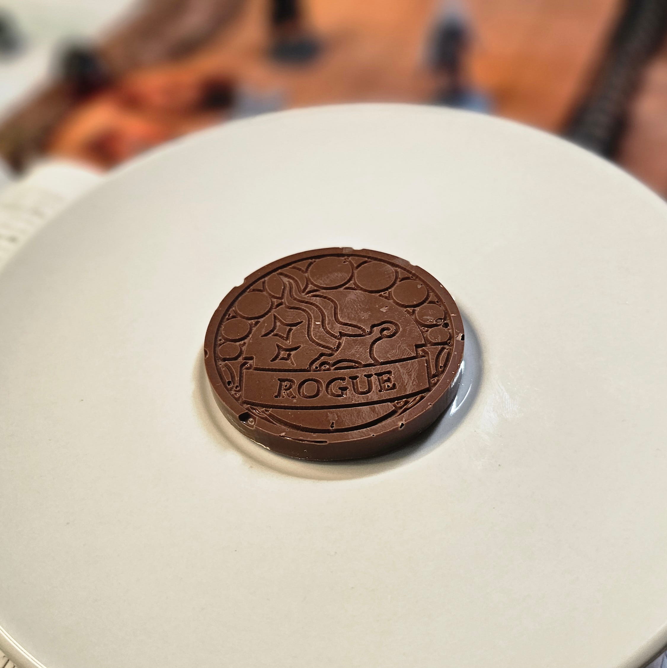 Home Milk chocolate crunch rogue inspiration coin displayed on a white plate with tabletop RPG miniatures, dice, and dungeon playmat blurred in the background.
