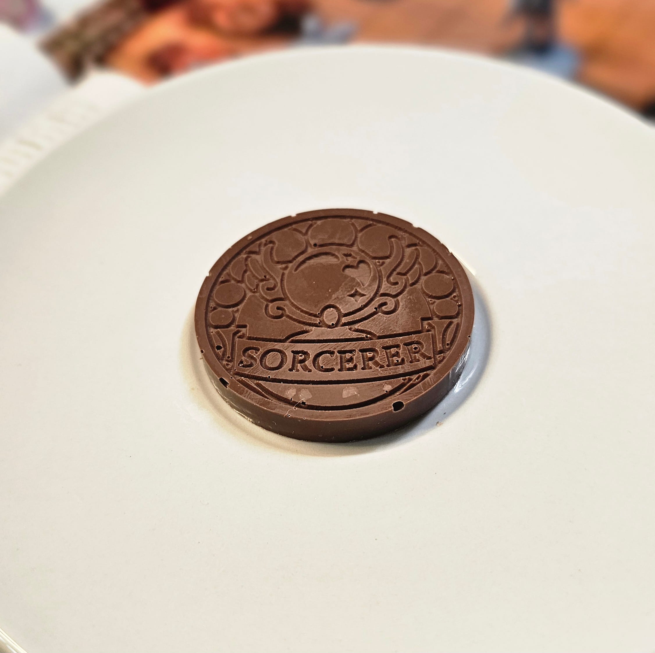 Home Milk chocolate crunch sorcerer inspiration coin displayed on a white plate with tabletop RPG miniatures, dice, and dungeon playmat blurred in the background.