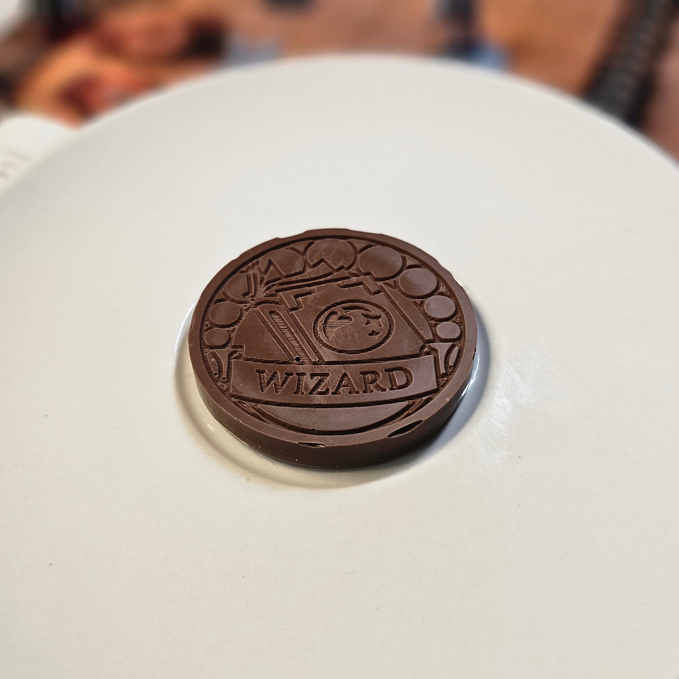 Home Milk chocolate crunch wizard inspiration coin displayed on a white plate with tabletop RPG miniatures, dice, and dungeon playmat blurred in the background.
