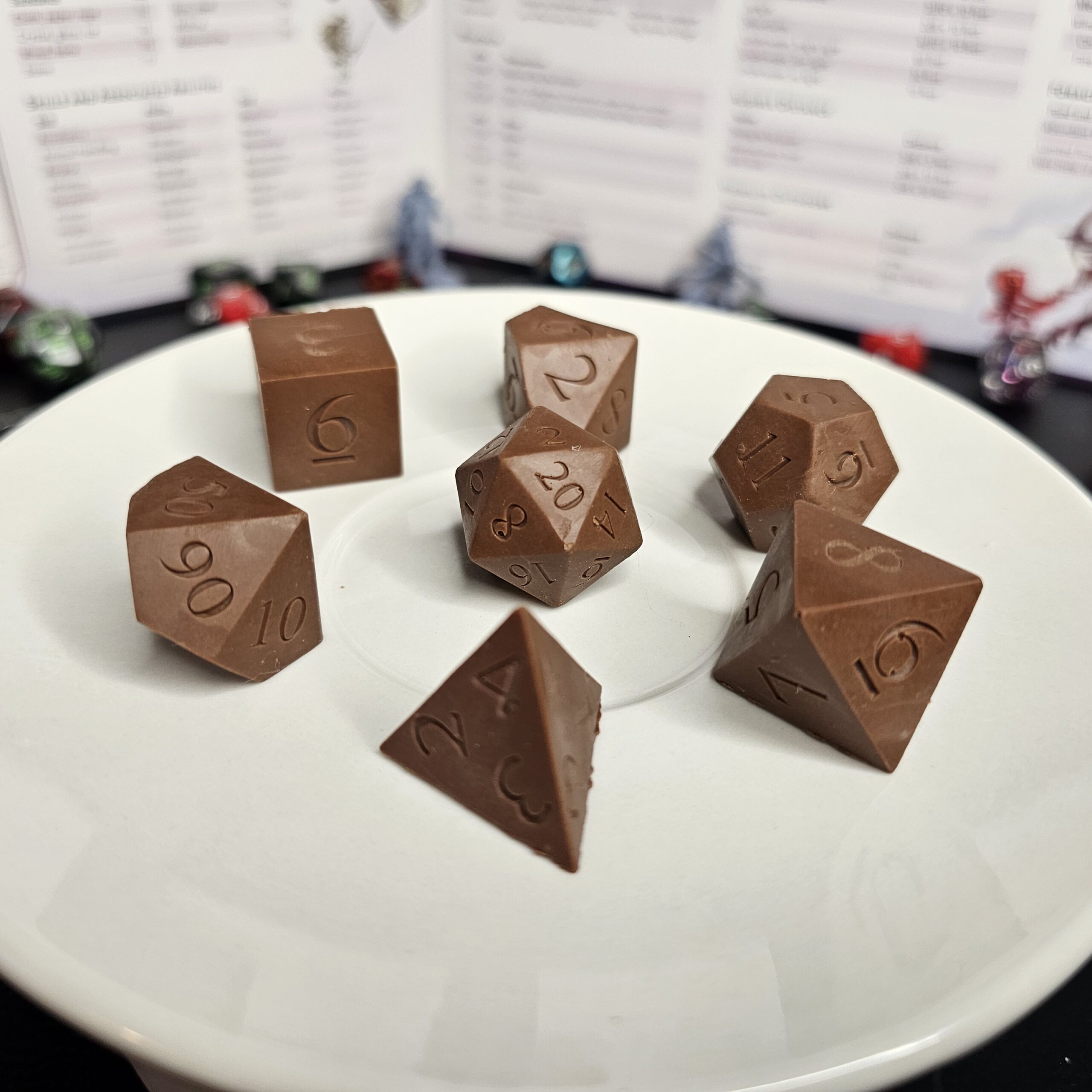 Strawberry Chocolate Dice Milk chocolate crunch D&D dice displayed on a white plate with tabletop RPG miniatures, dice, and a Dungeon Master screen blurred in the background.